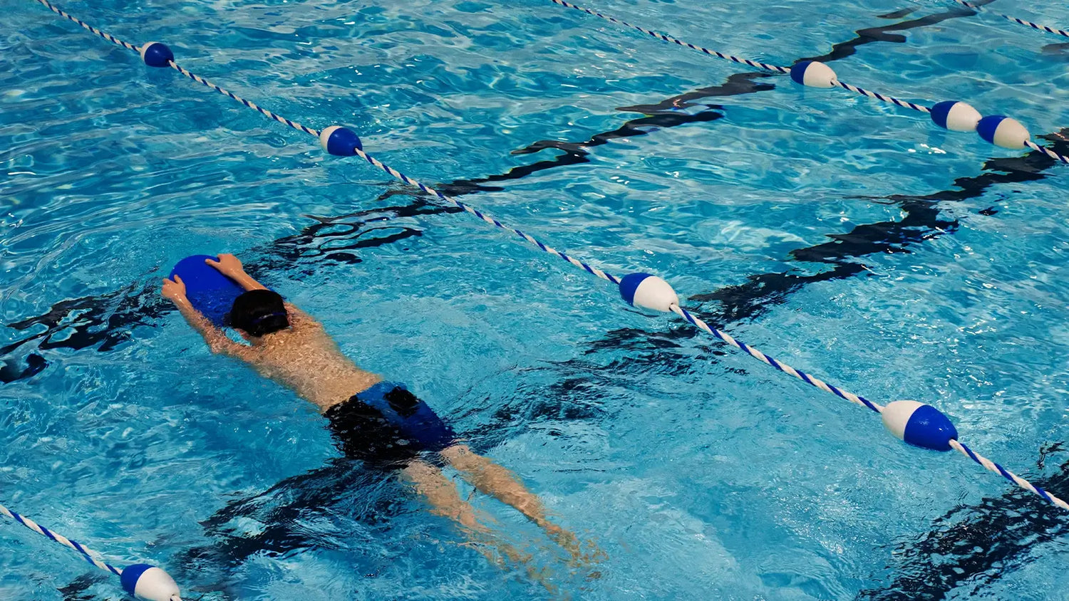 Beginner swimmer practicing with a blue kickboard between lane ropes in an indoor pool
