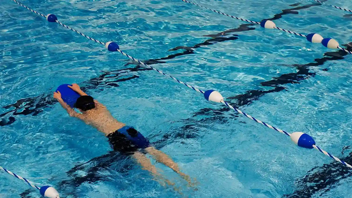Beginner swimmer practicing with a blue kickboard between lane ropes in an indoor pool